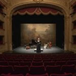 A wooden stage features a centered vintage microphone on a stand. A dark curtain forms the backdrop, illuminated by